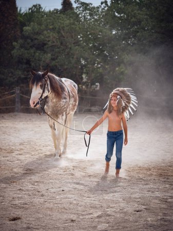 Foto de Joven chico delgado con sombreros sosteniendo riendas de caballo mientras pasea en tierra arenosa contra árboles verdes y mirando hacia otro lado - Imagen libre de derechos