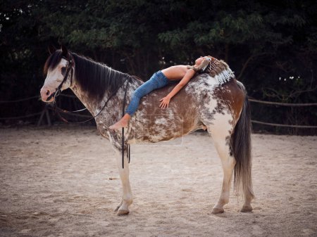 Foto de Vista lateral del niño joven con casco de plumas acostado a caballo mientras mira hacia arriba con la mano en el vientre del caballo en el paddock - Imagen libre de derechos