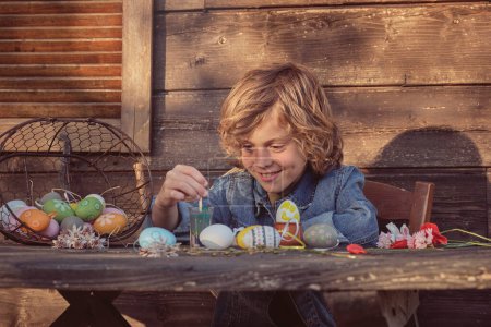 Téléchargez les photos : Petit enfant mignon peignant des œufs pour Pâques à la table en bois le jour ensoleillé - en image libre de droit