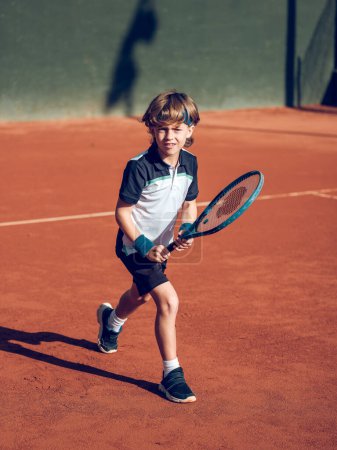 Full bode of focused cute preteen boy in sportswear playing tennis with racket on hard court