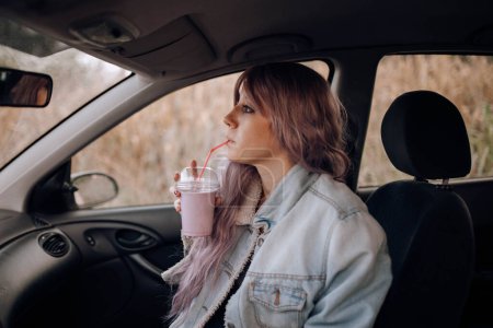 Photo for Side view of young modern woman with pink hairstyle drinking smoothie from plastic cup with straw sitting in car - Royalty Free Image