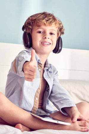 Photo for Positive barefoot boy sitting with crossed legs and enjoying songs from tablet in wireless headphones and showing like sign - Royalty Free Image
