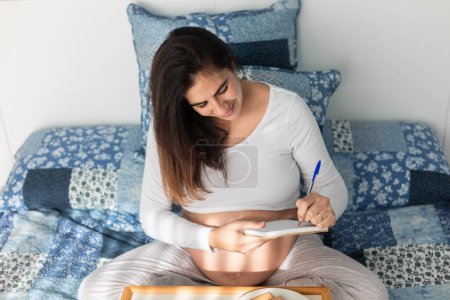 Téléchargez les photos : D'en haut de la femelle enceinte assise sur le lit et écrivant dans un cahier pendant le petit déjeuner le matin - en image libre de droit