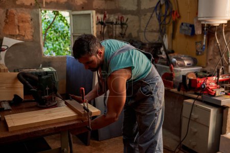 Foto de Vista lateral de carpintero masculino experto instalando abrazadera en tablón de madera mientras trabaja en carpintería ligera profesional con varios equipos - Imagen libre de derechos