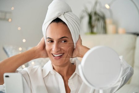 Téléchargez les photos : Femme adulte gaie avec un visage parfait propre et avec une serviette sur la tête souriante et regardant le miroir tout en touchant la tête et en enregistrant vlog sur smartphone après la douche - en image libre de droit