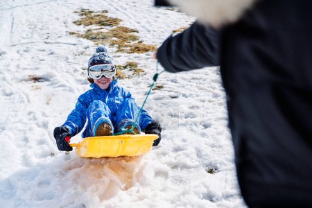 Photo for Crop parent pulling plastic sledge with gleeful son in outerwear with goggles and hat on cold winter day in nature - Royalty Free Image