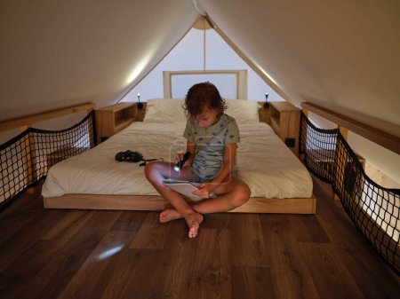 Photo for Full body of barefoot focused boy with glowing flashlight reading notepad while sitting on white bed in mansard at home - Royalty Free Image