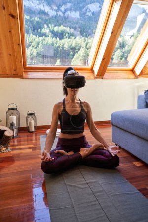 Photo for Full body of barefoot woman in VR headset meditating in Padmasana yoga asana while sitting with crossed legs on mat on wooden floor in mansard bedroom - Royalty Free Image
