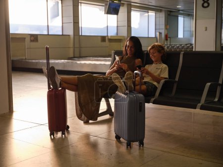 Téléchargez les photos : Corps complet de la mère et du fils assis dans des fauteuils avec les jambes sur les valises en attendant le vol à l'aéroport et en utilisant des smartphones - en image libre de droit