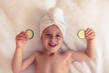 Téléchargez les photos : Vue de dessus du petit enfant souriant avec serviette sur la tête reposant sur le lit et tenant des tranches de concombre tout en regardant la caméra - en image libre de droit