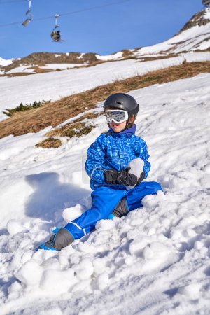 Photo for Kid in outerwear with goggles and helmet holding snow lump and looking away while sitting on slope on cloudless day on ski resort - Royalty Free Image