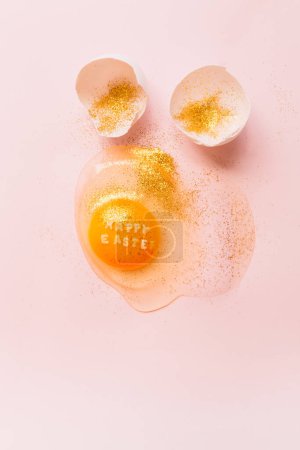 Photo for From above raw broken egg with text Happy Easter on yellow yolk covered in glitter and broken eggshells on pink background - Royalty Free Image