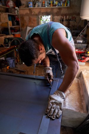 Téléchargez les photos : Artisanat masculin anonyme portant des gants de protection debout à l'établi et faisant de la métallurgie dans le garage - en image libre de droit