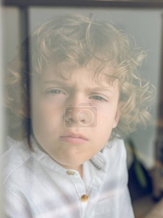 Foto de A través de vidrio de chico lindo mirando a la cámara con cara seria mientras está de pie cerca de la ventana en la sala de luz en casa - Imagen libre de derechos
