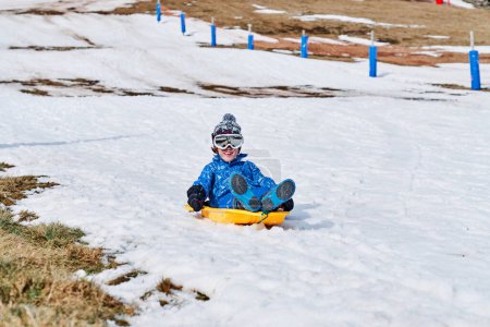 Photo for Boy in outerwear with goggles and hat smiling cheerfully and riding plastic sleigh on snowy slope in weekend on ski resort - Royalty Free Image