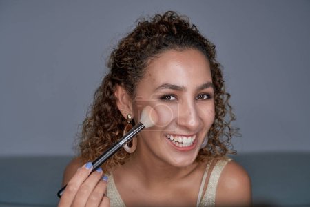 Photo for Delighted curly haired female visagiste applying glowing highlighter powder on cheek with professional brush and looking at camera - Royalty Free Image