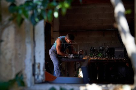 Téléchargez les photos : Vue latérale du menuisier masculin en tenue de travail debout sur l'établi en train de couper du métal à l'aide d'un puzzle électrique professionnel à la menuiserie - en image libre de droit
