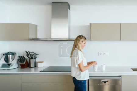 Foto de Vista lateral de la mujer de pie en el mostrador de la cocina con varios utensilios, mientras que la fabricación de té en tazas blancas en la luz apartamento moderno - Imagen libre de derechos