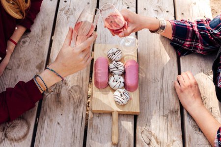 Téléchargez les photos : D'en haut les mains des femmes avec des verres à champagne assis à la table de pique-nique avec de savoureux beignets sur une planche en bois - en image libre de droit
