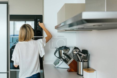 Foto de Vista trasera de la mujer anónima en traje casual que enciende el moderno horno de microondas negro mientras está de pie en la cocina con utensilios de cocina modernos - Imagen libre de derechos