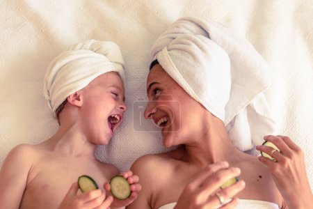 Photo for Top view of cheerful woman and kid with towels on heads resting on white blanket holding cucumber slices and looking at each other - Royalty Free Image