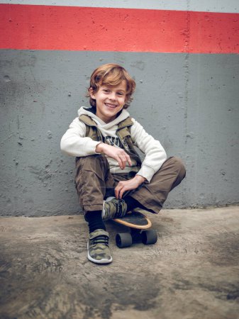 Téléchargez les photos : Corps complet d'un garçon élégant assis sur une planche à roulettes sur un sol en béton et appuyé sur un mur tout en souriant et en regardant la caméra - en image libre de droit