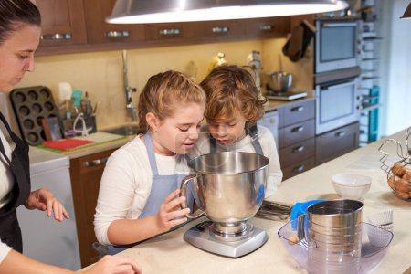 Photo for From above of curious little sister and brother with blond hair in aprons looking into metal bowl while standing at counter and baking pastry with crop young mother in kitchen - Royalty Free Image