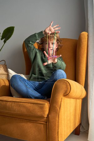 Photo for Positive preteen curly haired boy in casual clothes sitting in armchair with crossed legs and playing game while looking at camera through multicoloured string - Royalty Free Image