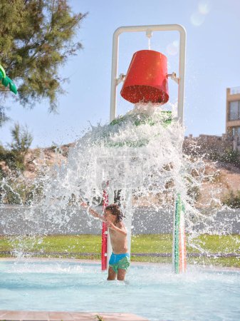 Téléchargez les photos : Petit garçon debout dans la piscine avec éclaboussures d'eau d'en haut pendant la journée ensoleillée - en image libre de droit