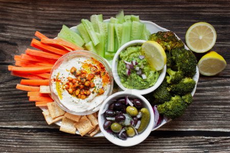 Photo for From above appetizing healthy hummus green sauce cut carrot cucumbers and green broccoli salted cucumbers decorated with sliced lemon in white bowls on wooden table - Royalty Free Image