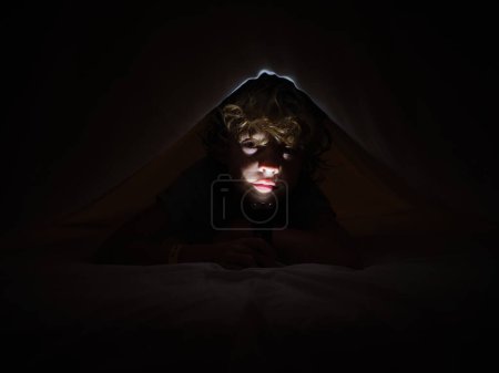 Téléchargez les photos : Mignon garçon éclairant visage avec une lampe de poche lumineuse tout en étant couché sur un lit confortable sous la couverture dans la chambre noire la nuit - en image libre de droit
