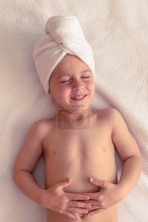 Foto de Vista superior de un niño pequeño encantado con el torso desnudo y toalla en la cabeza acostado sobre una manta blanca con los ojos cerrados - Imagen libre de derechos