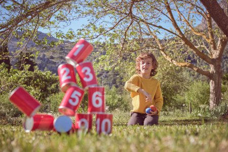 Foto de Chico positivo arrodillado en prado herboso y lanzando bolsas en pirámide de latas rojas mientras juega juego en la naturaleza - Imagen libre de derechos
