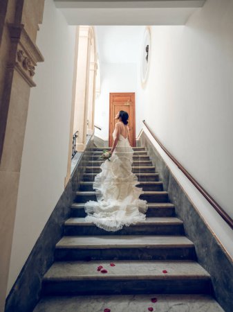 Téléchargez les photos : De dessous de vue arrière mariée méconnaissable en robe de mariée blanche avec volants et dentelle marchant à l'étage sur des marches avec des pétales de rose rouge et regardant la fenêtre de rêve - en image libre de droit
