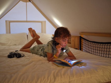 Photo for Full body of focused boy reading book with shining flashlight while lying on comfortable white bed with binocular on mansard - Royalty Free Image