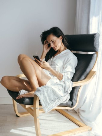 Photo for Full body side view of charming female with bare legs text messaging on cellphone while sitting in armchair near window - Royalty Free Image