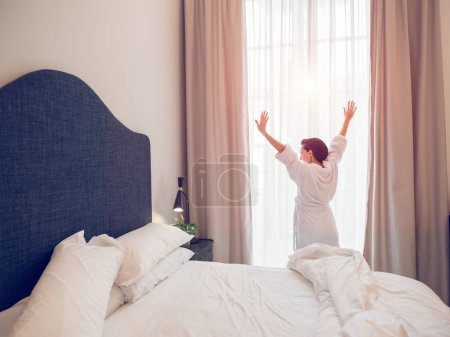 Téléchargez les photos : Vue arrière de la femme méconnaissable en peignoir blanc étirant les bras vers le haut tout en se tenant près de la fenêtre après s'être réveillée dans la chambre d'hôtel à l'heure du matin - en image libre de droit