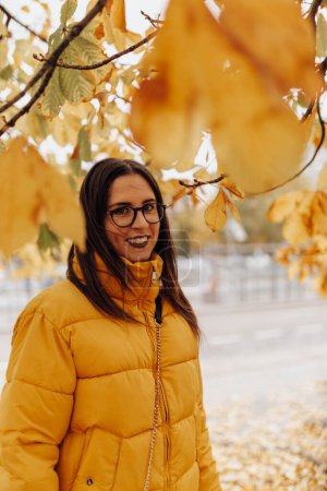 Photo for Beautiful young lady in stylish warm clothes smiling and standing on tree on amazing autumn day on city street - Royalty Free Image