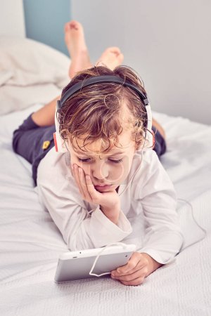 Foto de Alto ángulo de chico descalzo en auriculares acostado en la cama y viendo dibujos animados en la tableta mientras se apoya en la mano - Imagen libre de derechos