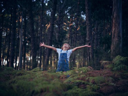 Photo for Cute boy in denim overall with spread arms standing in woods with green fern plants and trees on summer day - Royalty Free Image