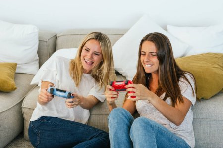 Téléchargez les photos : Recadrage d'amies positives avec des contrôleurs de jeu jouant au jeu vidéo assis sur le sol appuyé sur le canapé dans le salon à la maison - en image libre de droit
