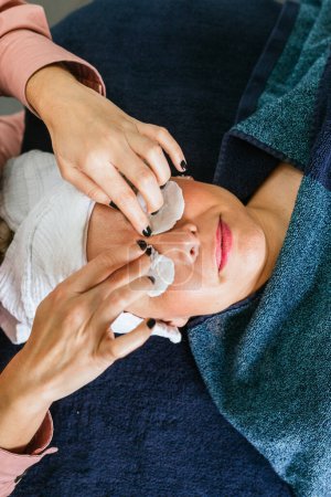 Photo for Top view of crop unrecognizable cosmetician using cotton pads and applying tonic on eyes of female customer lying on table in beauty salon during skincare procedure - Royalty Free Image