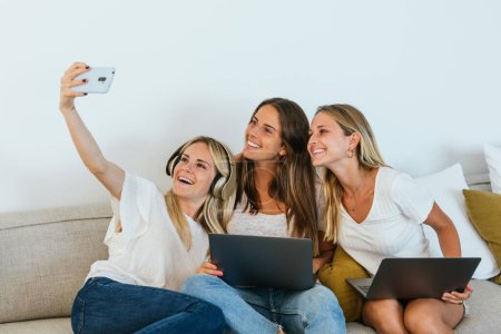 Foto de Mujer tomando una selfie con el teléfono celular a alegres amigas sentadas en el sofá en la sala de estar - Imagen libre de derechos