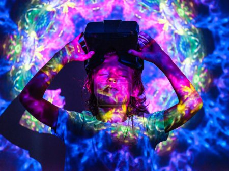 Foto de Niño con los ojos cerrados y auriculares VR en la cabeza de pie bajo neón iluminación proyector abstracto colorido - Imagen libre de derechos