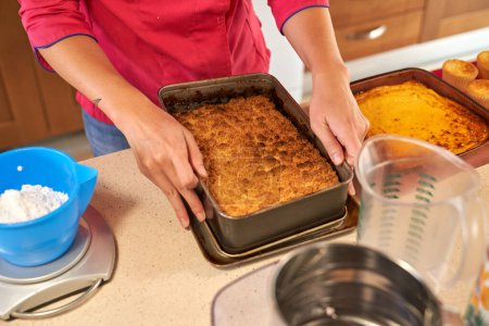 Téléchargez les photos : Recadrer confiseur professionnel anonyme sortir tarte de moule en métal sur le comptoir de cuisine avec divers ustensiles de cuisine et muffins à la maison - en image libre de droit