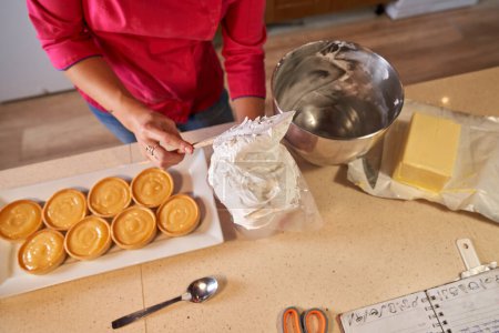 Foto de Vista superior de la chef femenina de cosecha en bolsa de repostería de relleno uniforme con crema de mantequilla para decorar tartaletas en panadería casera - Imagen libre de derechos