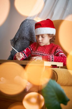 Foto de Niño enfocado en el sombrero de Santa sentado en un sillón cómodo y mirando hacia abajo mientras dibuja en la mesa - Imagen libre de derechos