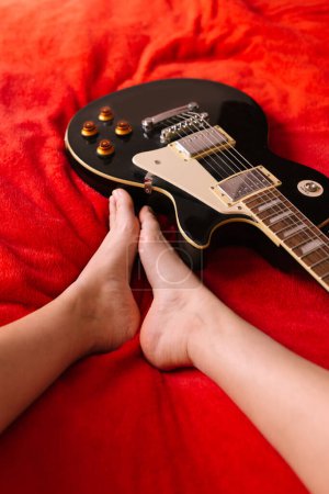 Téléchargez les photos : D'en haut du guitariste féminin assis pieds nus sur le lit avec un instrument de musique électrique à la maison - en image libre de droit