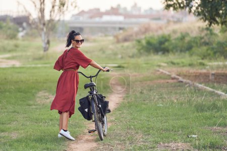 Foto de Vista lateral de la joven ciclista en maxi vestido rojo y gafas de sol caminando con bicicleta a lo largo del sendero mientras sonríe y mira por encima del hombro - Imagen libre de derechos
