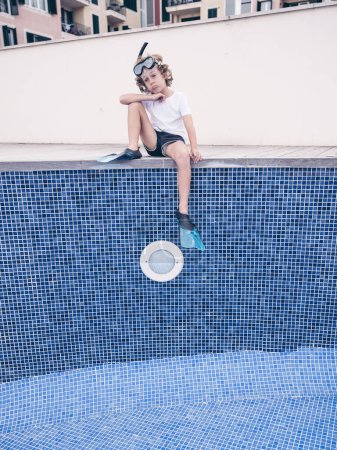Téléchargez les photos : De dessous de corps complet se demandant garçon préadolescent en masque de plongée avec tuba et palmes assis avec jambe courbée sur le côté de la piscine vide - en image libre de droit